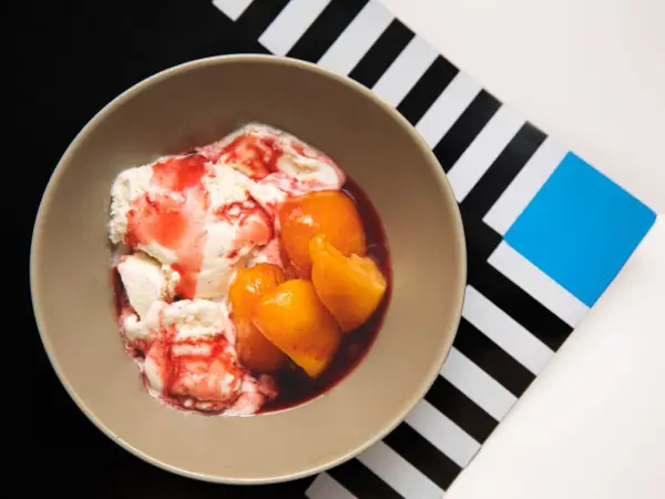En skål med krämig vaniljglass, saftiga persikohalvor och en röd bärsås. En klassisk peach melba som ser utsökt ut.