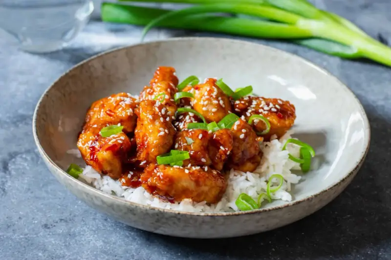 Krispig koreansk kyckling med sesamfrön och salladslök serveras på en bädd av vitt ris i en skål.