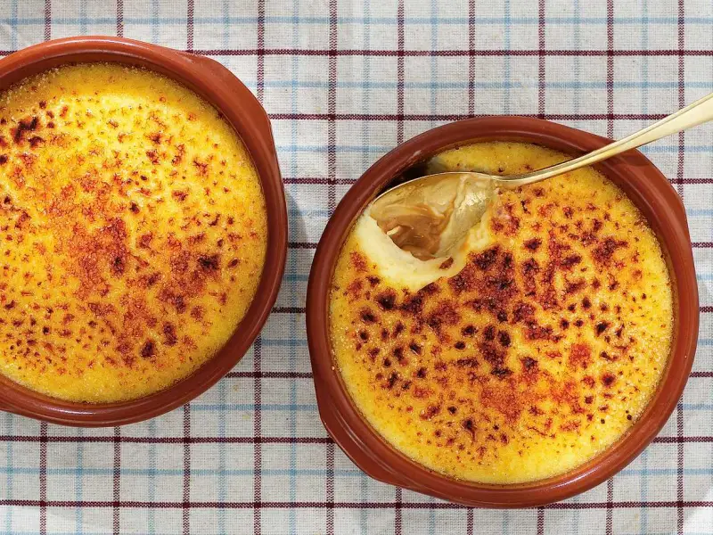 Två krämiga creme brulee i bruna skålar, en med en gyllene sked som lyfter fram den karamelliserade ytan.